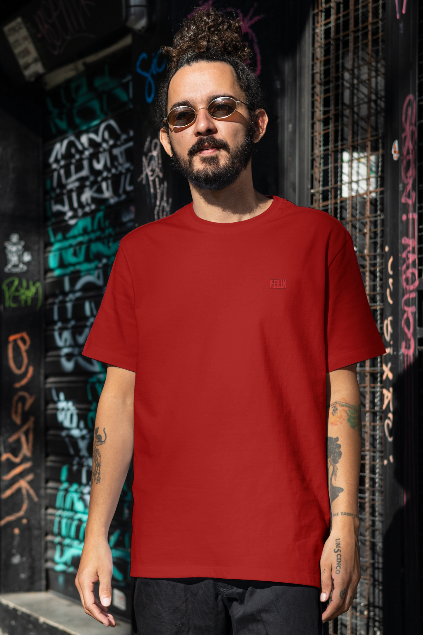Man wearing a red t-shirt and sunglasses standing in front of a graffiti-covered wall.