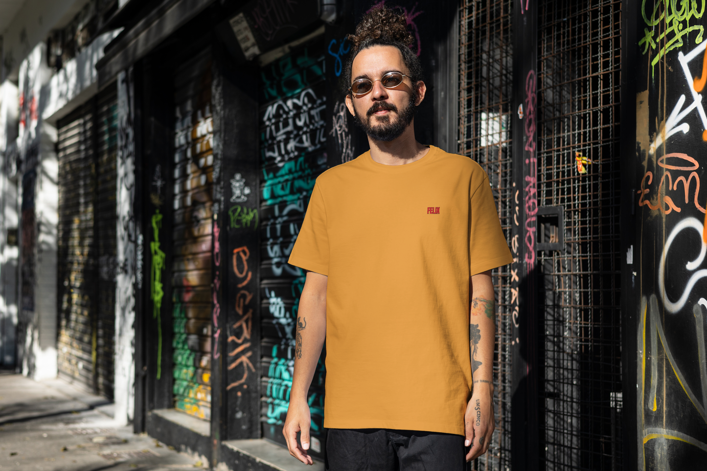 Man wearing a mustard yellow t-shirt with a small red logo, standing in front of a graffiti-covered wall.